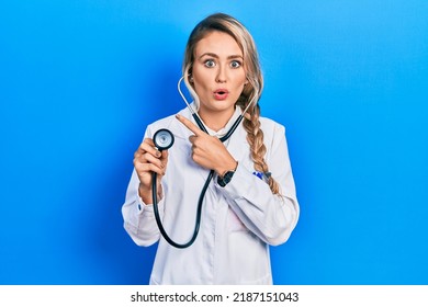 Beautiful young blonde doctor woman holding stethoscope surprised pointing with finger to the side, open mouth amazed expression. 