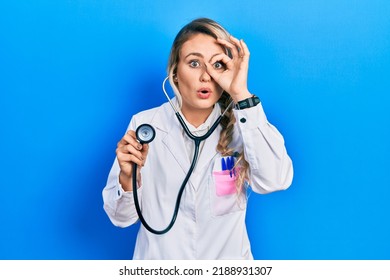 Beautiful young blonde doctor woman holding stethoscope doing ok gesture shocked with surprised face, eye looking through fingers. unbelieving expression. 