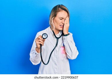 Beautiful Young Blonde Doctor Woman Holding Stethoscope Yawning Tired Covering Half Face, Eye And Mouth With Hand. Face Hurts In Pain. 