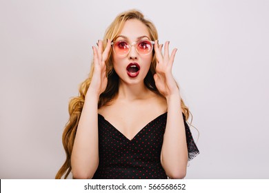 Beautiful young blonde girl with surprised face, young woman with opened mouth. Studio shot. Wearing pink cool glasses, black dress, has nice curly hair. Isolated background.