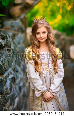 Beautiful young blonde princess in gold medieval dress and little crown. Pretty queen posing near stone castle.Warm art work.