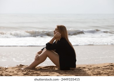 Beautiful young blonde woman in black shirt sits on the shore of the beach sad and depressed. The woman is pensive and looks at the ground exhausted and tired. The woman is suffering. - Powered by Shutterstock