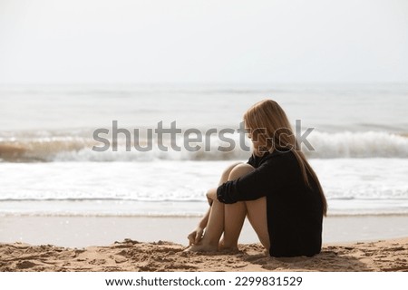 Beautiful young blonde woman in black shirt sits on the shore of the beach sad and depressed. The woman is pensive and looks at the ground exhausted and tired. The woman is suffering.