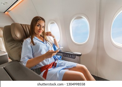 Beautiful Young Blonde Woman In Blue Dress Sits In Business Class Chair In Plane Cabin During Flight Near Porthole, Holding Her Smartphone 