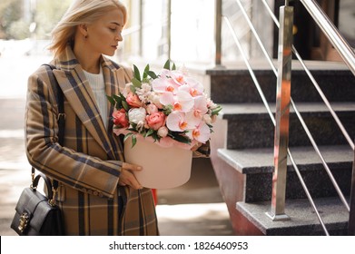 beautiful young blonde woman in coat holds pink round box with flower arrangement of roses and orchids.