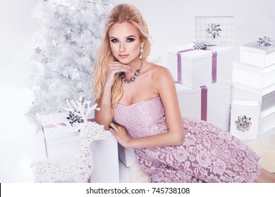 Beautiful Young Blonde Woman In Elegant Pink Dress Sitting Next To White Christmas Tree And Presents. Christmas Glamour Photo