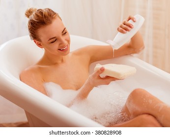 Beautiful young blonde woman enjoying pleasant bath with foam, putting shower gel on a wisp of bast and smiling