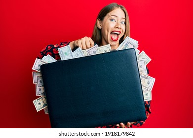 Beautiful young blonde woman holding briefcase full of dollars celebrating crazy and amazed for success with open eyes screaming excited. 