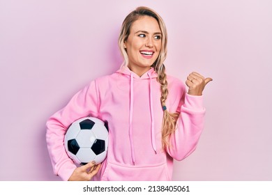 Beautiful young blonde woman holding soccer ball pointing thumb up to the side smiling happy with open mouth 