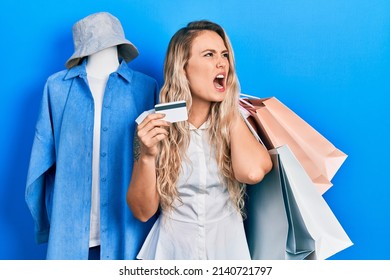 Beautiful Young Blonde Woman Holding Shopping Bags And Credit Card Angry And Mad Screaming Frustrated And Furious, Shouting With Anger. Rage And Aggressive Concept. 