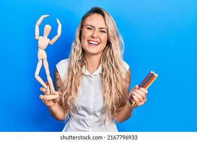 Beautiful young blonde woman holding wooden manikin and colored pencils smiling and laughing hard out loud because funny crazy joke. 