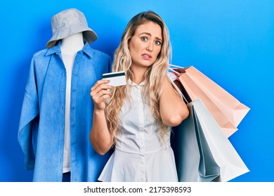 Beautiful Young Blonde Woman Holding Shopping Bags And Credit Card Clueless And Confused Expression. Doubt Concept. 