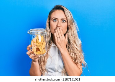 Beautiful young blonde woman holding dry spaghetti covering mouth with hand, shocked and afraid for mistake. surprised expression 