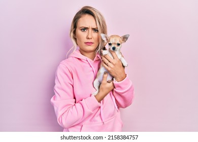 Beautiful young blonde woman hugging cute chihuahua dog skeptic and nervous, frowning upset because of problem. negative person. 