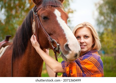 A Beautiful Young Blonde Woman Hugs Her Horse And Looks Into The Camera. The Concept Of Love For Nature, Human Interaction With Nature And Animals.