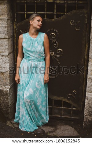 A beautiful young blonde woman in a long green dress is standing at a metal door. Vintage photo of a lady in a dress in a dungeon. Hopeless sadness, lack of freedom.