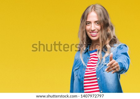 Beautiful young blonde woman over isolated background smiling friendly offering handshake as greeting and welcoming. Successful business.