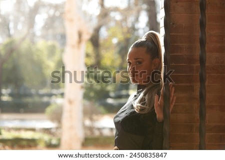 Beautiful young blonde woman with a ponytail in her hair leaning against a red brick wall and holding on to a fence. In the background a famous Seville park and a backlight in the golden hour