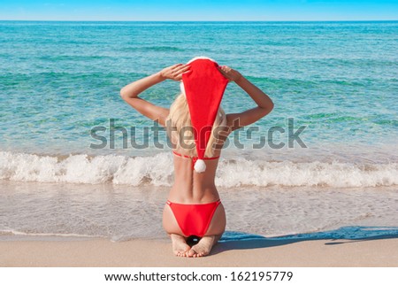 Beautiful young blonde woman in red christmas hat on sea beach. New year holidays in hot countries concept.