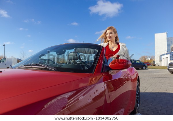 Beautiful young blonde woman in a red\
dress posing in a red car in the city on a sunny\
day