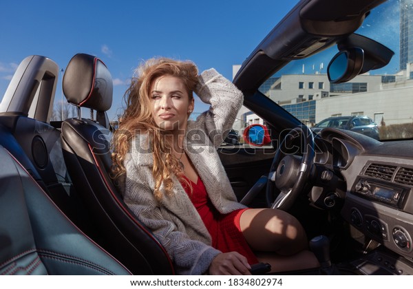 Beautiful young blonde woman in a red\
dress posing in a red car in the city on a sunny\
day