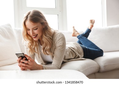 Beautiful Young Blonde Woman Relaxing On A Couch At Home, Using Mobile Phone