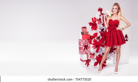 Beautiful Young Blonde Woman In Sexy Red Dress Standing Next To White Christmas Tree And Presents. Christmas Glamour Photo. A Lot Of Gifts. 