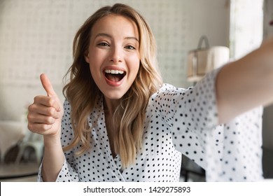 Beautiful young blonde woman sitting at the cafe indoors, taking a selfie, thumbs up