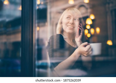 Beautiful Young Blonde Woman Sitting In A Cafe Talking On A Mobile Phone By The Window With A Reflection Of Street