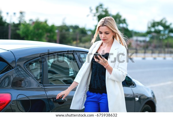 Beautiful young blonde woman speaking speaking on\
phone near black car.