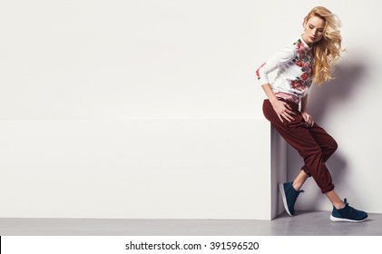 Beautiful Young Blonde Woman In A Sweatshirt Flower With Pattern And Jeans Posing In Studio. Fashion Photo