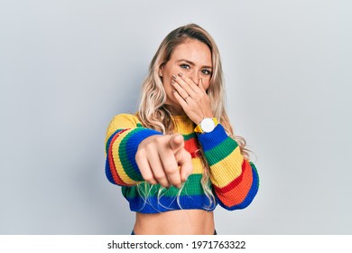 Beautiful Young Blonde Woman Wearing Colored Sweater Laughing At You, Pointing Finger To The Camera With Hand Over Mouth, Shame Expression 