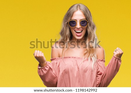 Beautiful young blonde woman wearing retro sunglasses over isolated background celebrating surprised and amazed for success with arms raised and open eyes. Winner concept.