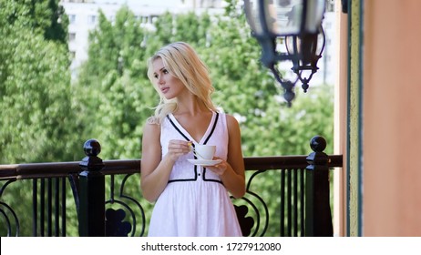 Beautiful young blonde woman in white dress drinking coffee, standing on the balcony on summer day in luxury hotel.