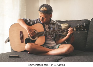 Beautiful Young Boy Playing Acoustic Guitar. Music Student Exercises Alone At Home. Trendy Caucasian Child Plays Instruments Sitting Legs Crossed On The Sofa. Blonde Guy Enjoying Melody Of A Song