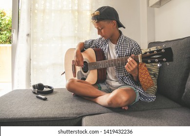 Beautiful Young Boy Playing Acoustic Guitar. Music Student Exercises Alone At Home. Trendy Caucasian Child Plays Instruments Sitting Legs Crossed On The Sofa. Blonde Guy Enjoying Melody Of A Song