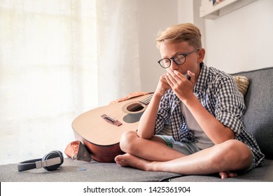 Beautiful young boy playing mouth harmonica. Music student exercises alone at home. Trendy caucasian child plays instruments sitting legs crossed on the sofa. Blonde guy doing musical homework - Powered by Shutterstock