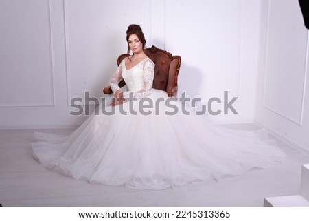 A beautiful young bride model in long lace dress sits on armchair in minimalist white studio interior. Wedding photography, portrait.
