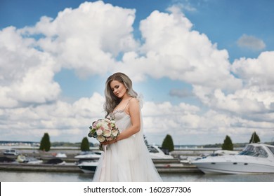 Beautiful Young Bride With Platinum Blond Hair In Lace Wedding Dress Keeps Bouquet Of Fresh Flowers And Posing On The Lake Coast. Young Woman With Wedding Hairstyle In Stylish Dress Posing Outdoors.