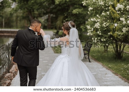 A beautiful young bride is walking with her groom in the summer park. Photo of the bride and groom from behind. Beautiful wedding white dress. Walks in the park. A happy and loving couple.