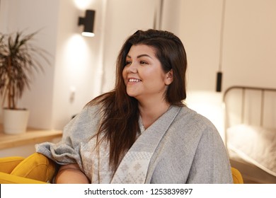 Beautiful Young Brunette Chubby Woman Posing Indoors, Looking Away With Positive Charming Smile, Spending Nice Time At Home, Wearing Gray Wide Scarf. Body Positivity, Joy And Happiness Concept