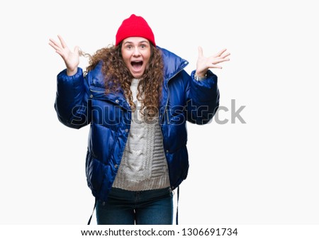 Beautiful young brunette curly hair girl wearing winter coat, wool cap and sweater over isolated background celebrating crazy and amazed for success with arms raised and open eyes screaming excited