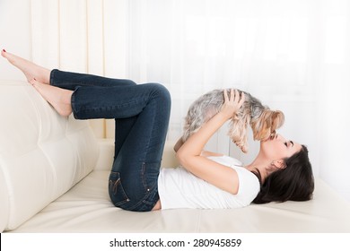 Beautiful young brunette girl playing with her yorkshire terrier at home laying down at sofa and kissing her pet - Powered by Shutterstock