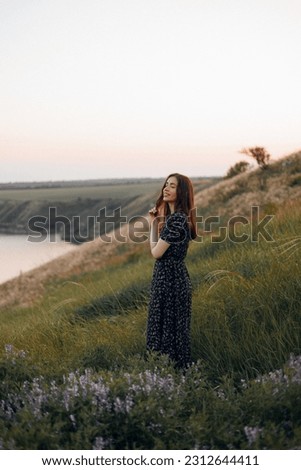 A beautiful young brunette girl stands among wildflowers and holds a flower sprig in her hands, looks away and smiles. summer cute portrait, pleasant face cheerful and sincere