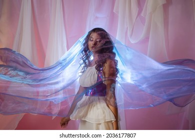 Beautiful Young Brunette In A Long White Dress, Light Flying Fabric, Silk Skirt, Portrait In A Fabulous Shimmer. Surreal Style, Translucent Fabrics On The Background