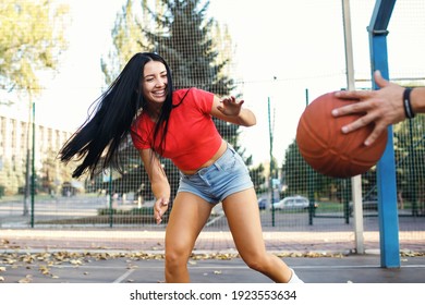 A Beautiful Young Brunette Is Playing Basketball With Her Boyfriend And Is Trying To Pick Up The Ball. The Sports Couple Is Having Fun And Enjoying The Process. Girl In Denim Shorts And A Red T-shirt