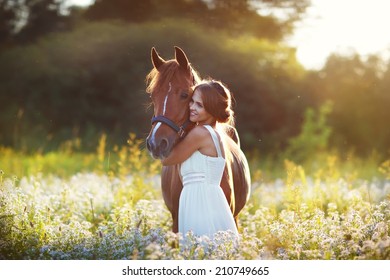  A beautiful young brunette smiling woman in a white dress embracing a horse in a sunny summer day. Adults and nature.