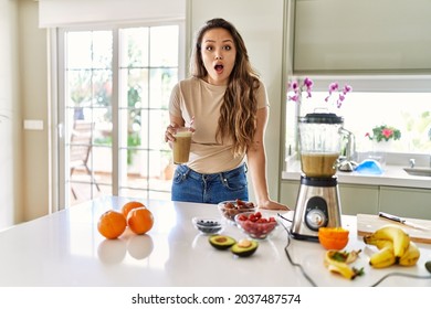 Beautiful Young Brunette Woman Drinking Glass Of Smoothie At The Kitchen Scared And Amazed With Open Mouth For Surprise, Disbelief Face 