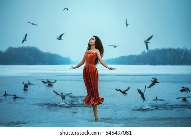 Beautiful young brunette woman flies with the birds. Background shore of a frozen river. . Sad face with a glimmer of hope to escape from the routine of everyday life. Fantasy photo, creative color