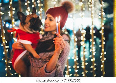 Beautiful young brunette woman with French bulldog enjoying Christmas or New Year night on a city street.  Selective focus on candles.